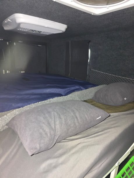 RV rental interior: Cozy sleeping area with grey pillows, blue bedding, and an overhead AC unit for comfortable travel.