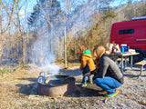 Enjoying an RV rental adventure! Family cooks by campfire, with their red RV at a wooded campsite.
