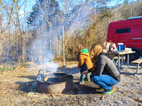 Enjoying an RV rental adventure! Family cooks by campfire, with their red RV at a wooded campsite.