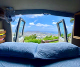Comfortable RV rental bed with open doors revealing a stunning panoramic city, mountain, and ocean view.