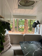 Cozy RV rental interior with gray sofa, black marble table, and a fan, looking out to a serene forest view.