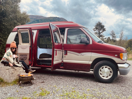 Enjoying a cozy moment by an RV rental: a person roasting marshmallows by a fire pit next to a camper van with mountains.