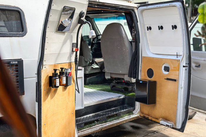  enjoying a game projected on a screen in her custom mobile living space.|This RV rental camper van features a clever outdoor shower with soap and smart storage for road trips.|A woman takes an outdoor shower from her custom camper van