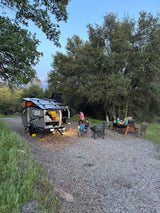 Family enjoying an RV rental. A grey "Cricket" trailer, picnic table, and chairs are set up at a gravel campsite surrounded by trees.
