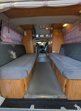 Looking into an RV rental's interior, featuring dual gray bench seats with wooden bases, a carpeted floor, and front cabin view.