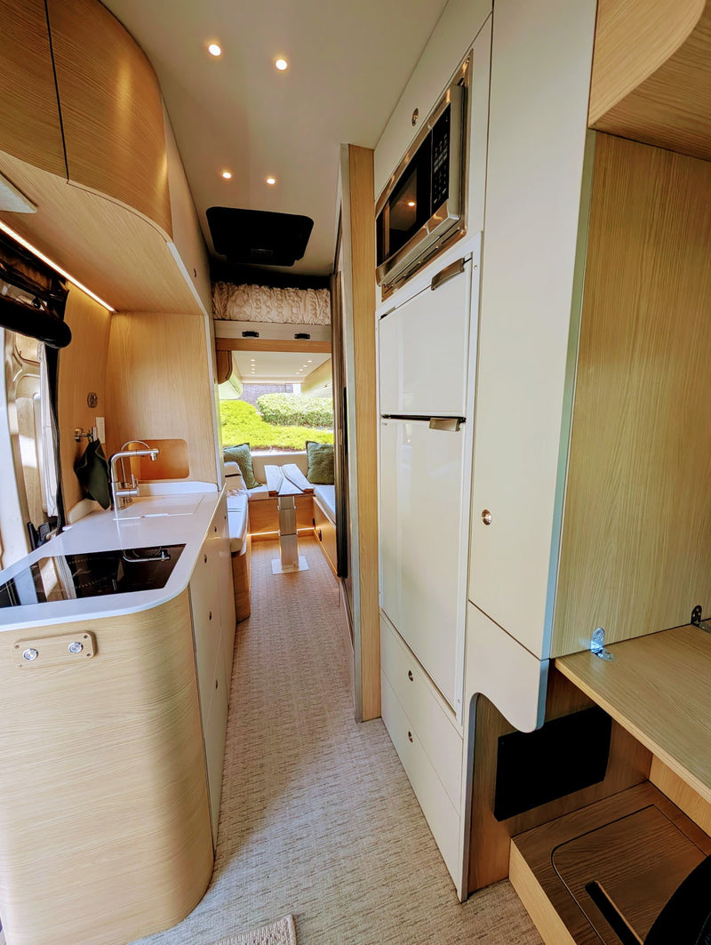 Bright RV rental interior: modern compact kitchen with sink/stove, microwave, fridge, and dinette leading to scenic views. — photo 2