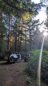 White off-road teardrop trailer at a forest campsite, sun rays piercing through. Perfect for an RV rental adventure.