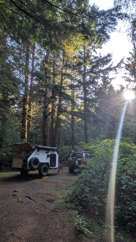 White off-road teardrop trailer at a forest campsite, sun rays piercing through. Perfect for an RV rental adventure.