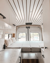 Modern RV rental interior: convertible bed, white and brown slatted ceiling, bright kitchen area.