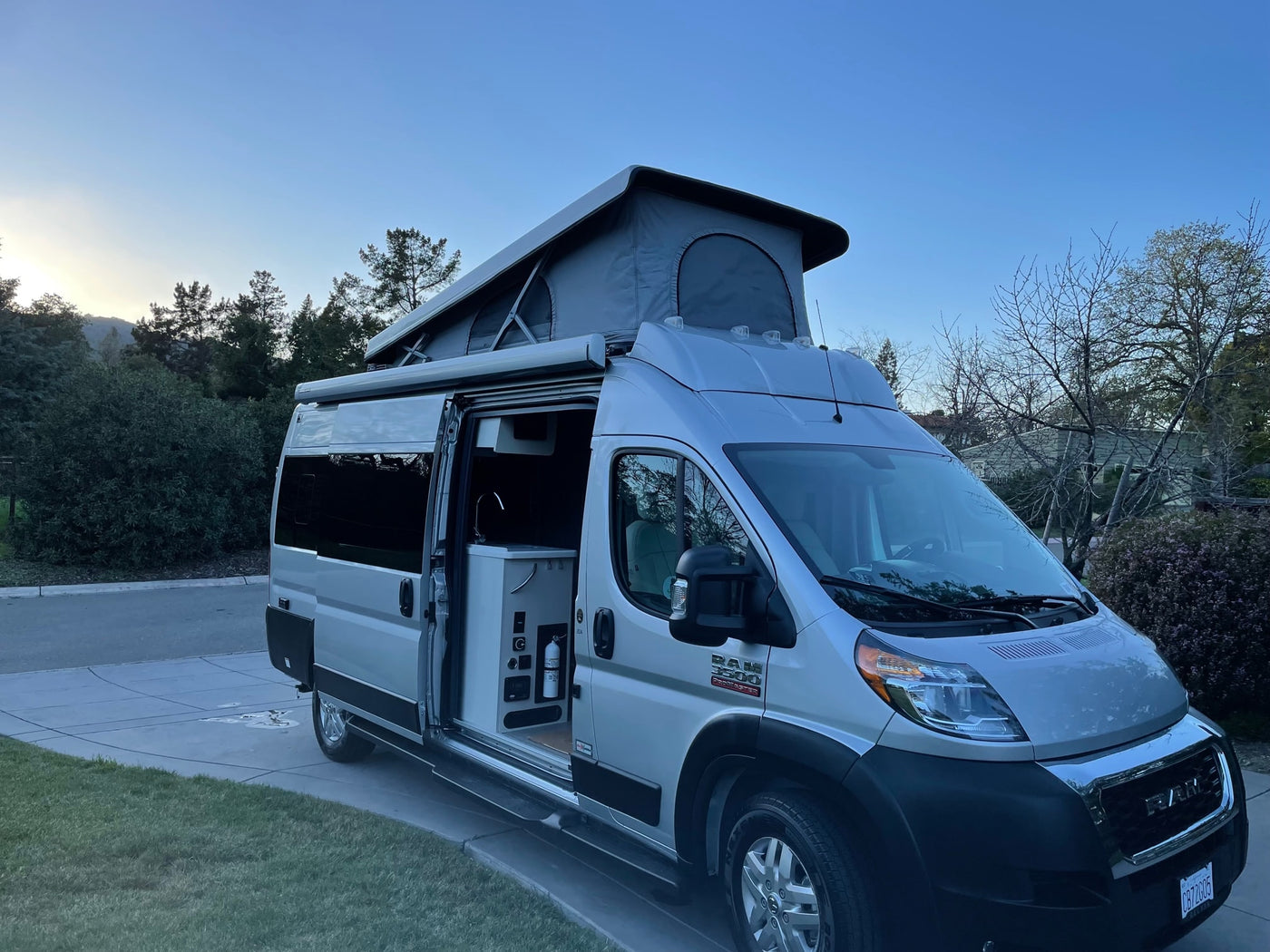 This silver Ram ProMaster pop-top camper van, side door open, is parked on a driveway. Consider it for your next rv rental! — photo 3