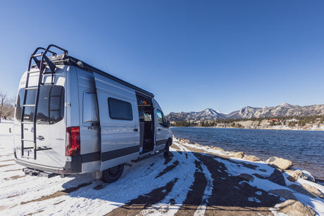 Silver RV rental by a tranquil lake with snow-dusted ground and majestic mountains under a clear blue sky.