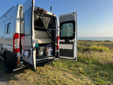 Silver RV rental camper van parked on a grassy cliff, back doors open revealing interior and ocean view. Enjoy scenic travels!