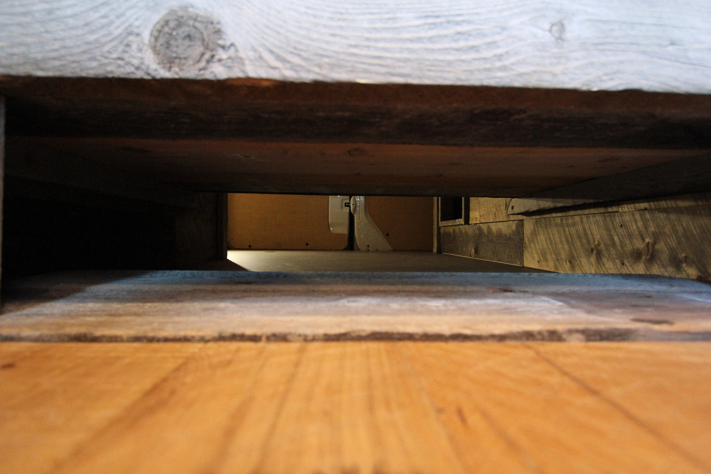 View from under a wooden bed in an RV rental, showing the interior floor and wall in the distance.