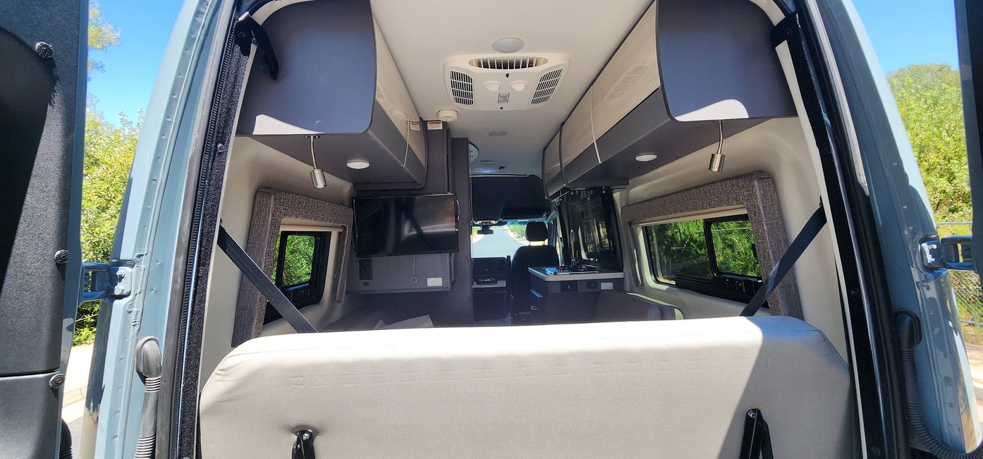 View from rear of a spacious RV rental. Interior shows overhead storage, TV, kitchenette, & driver's area.
