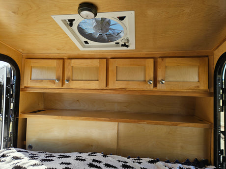 Cozy RV rental interior featuring natural wood ceilings, overhead storage cabinets, shelves, a ceiling fan, and a comfortable bed.