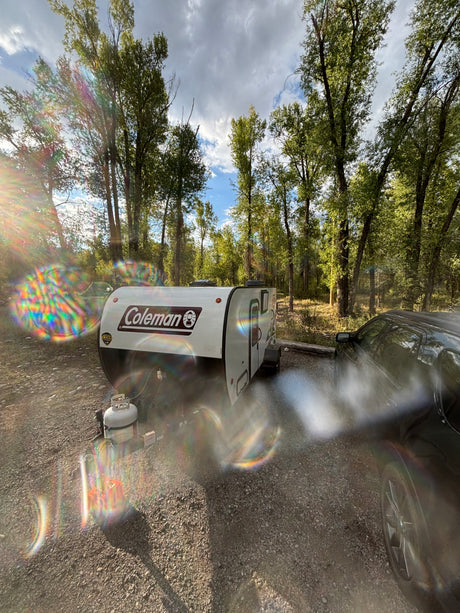 Coleman travel trailer RV rental and SUV at a bright, wooded campsite, with colorful lens flare.