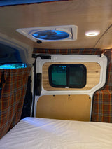 Inside an RV rental: a comfortable bed, plaid curtains, wood-paneled walls, and a ceiling fan with blue light.