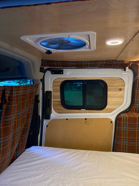 Inside an RV rental: a comfortable bed, plaid curtains, wood-paneled walls, and a ceiling fan with blue light.