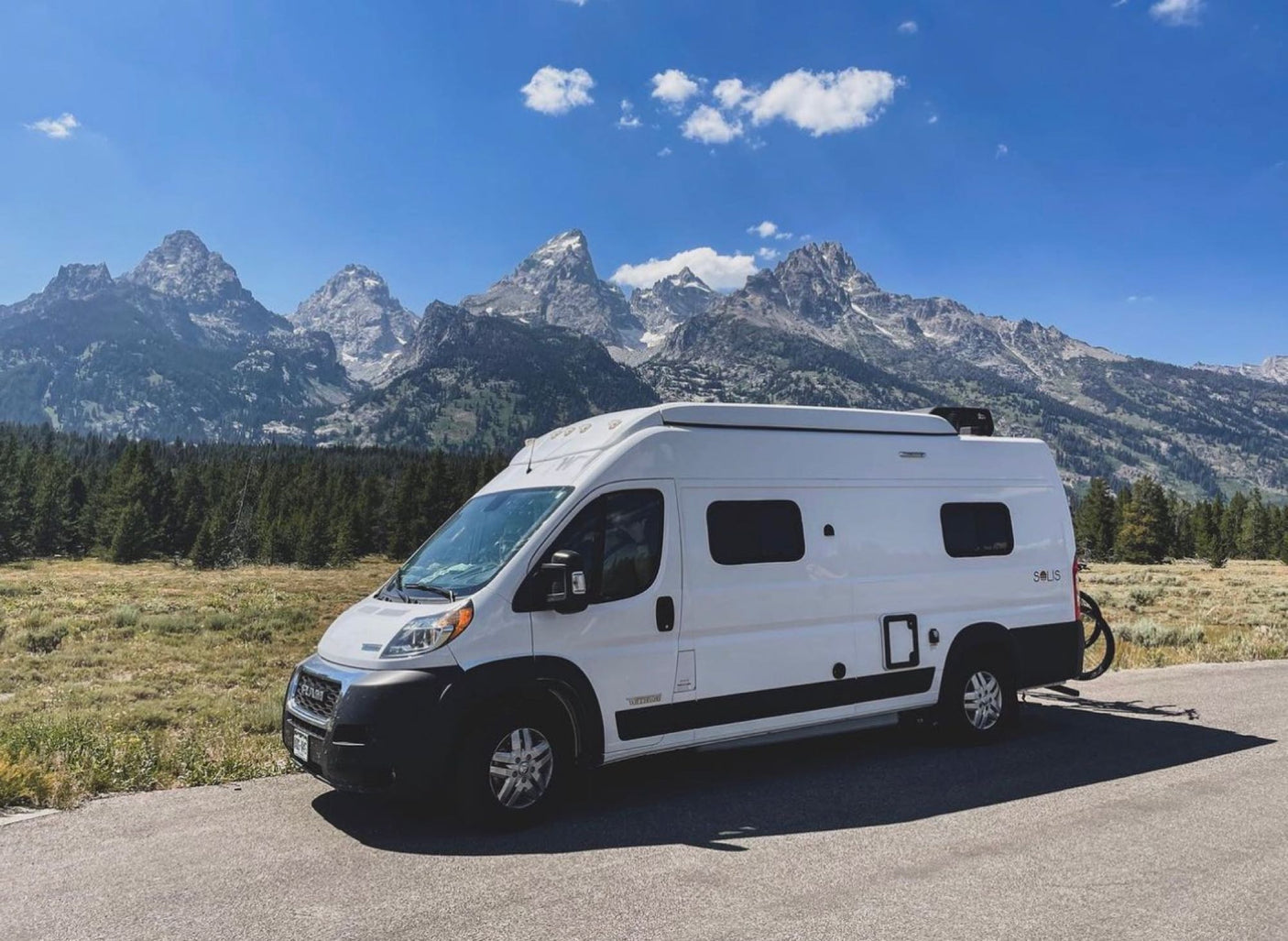 A white RV rental camper van parked on a scenic road with majestic Grand Teton mountains and green forests under a blue sky.