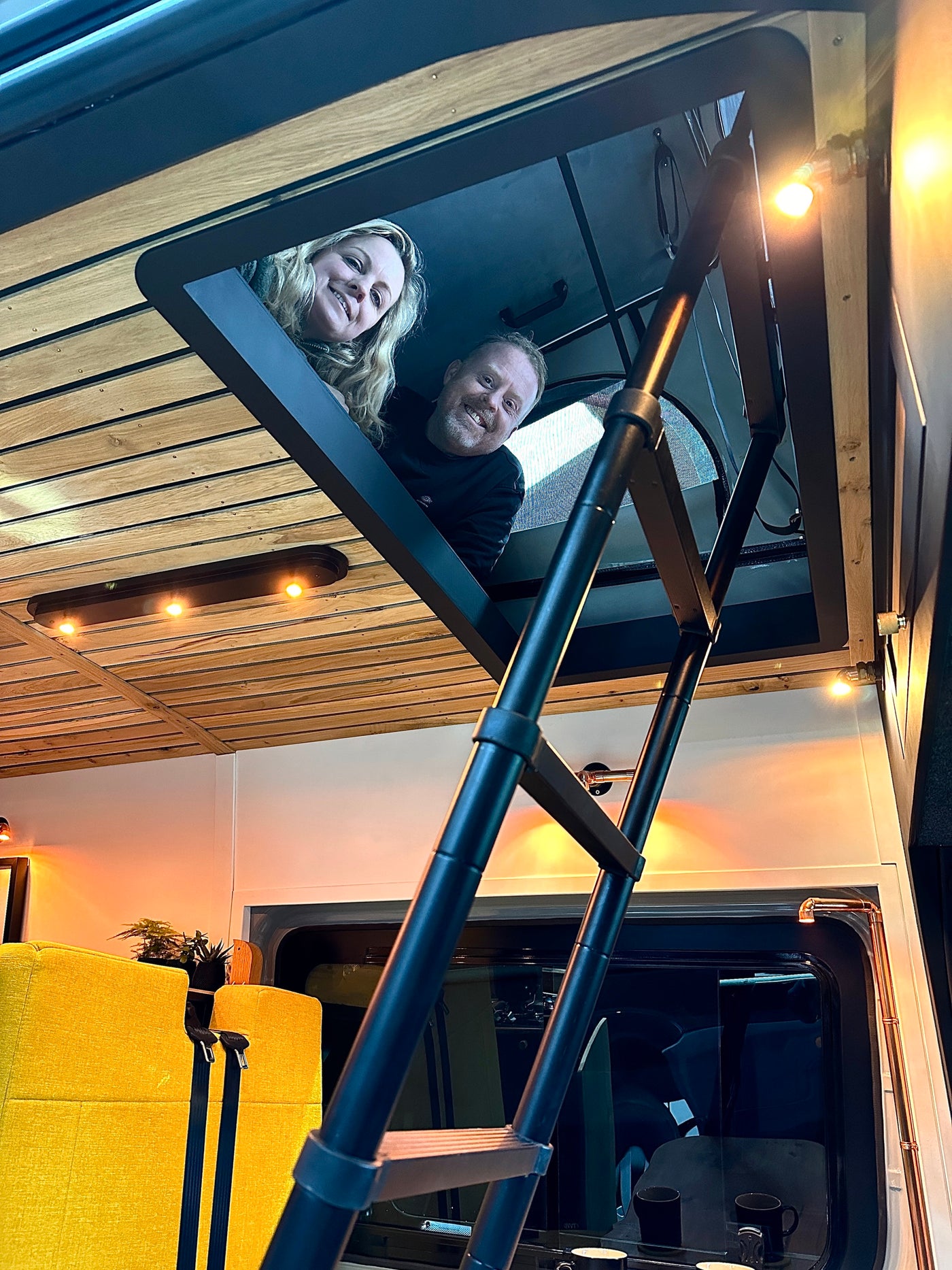 Smiling couple peeking from their RV rental loft. Cozy camper interior features a ladder, wood ceiling, & warm lighting. — photo 11