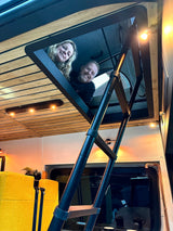 Smiling couple peeking from their RV rental loft. Cozy camper interior features a ladder, wood ceiling, & warm lighting.