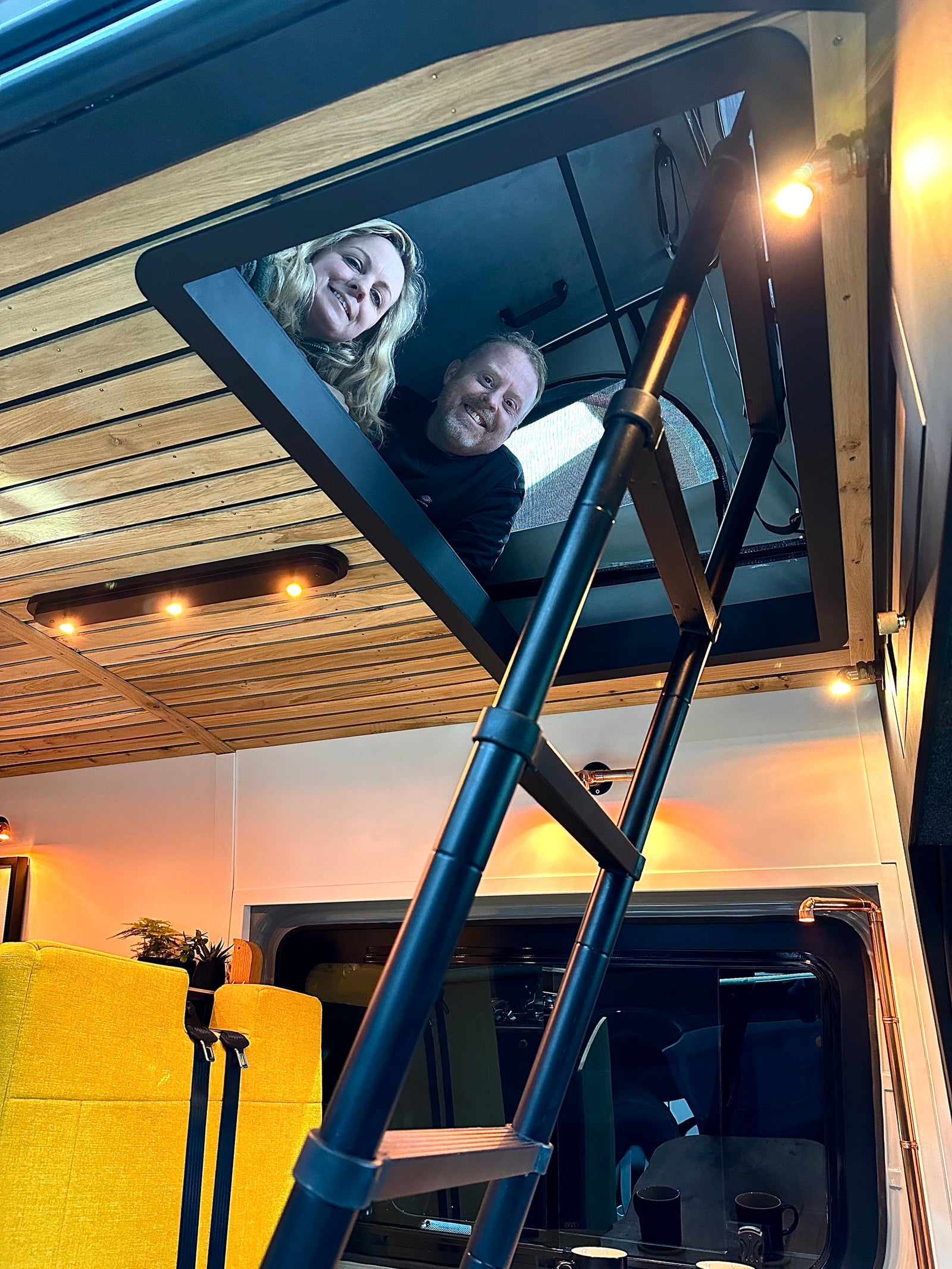 Smiling couple peeking from their RV rental loft. Cozy camper interior features a ladder, wood ceiling, & warm lighting.