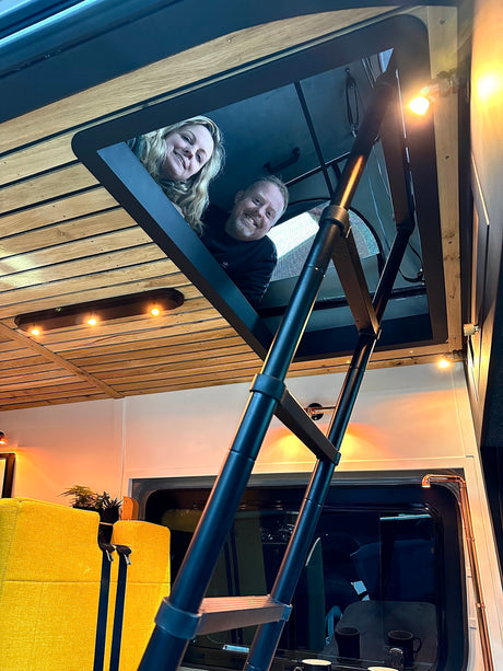 Smiling couple peeking from their RV rental loft. Cozy camper interior features a ladder, wood ceiling, & warm lighting.