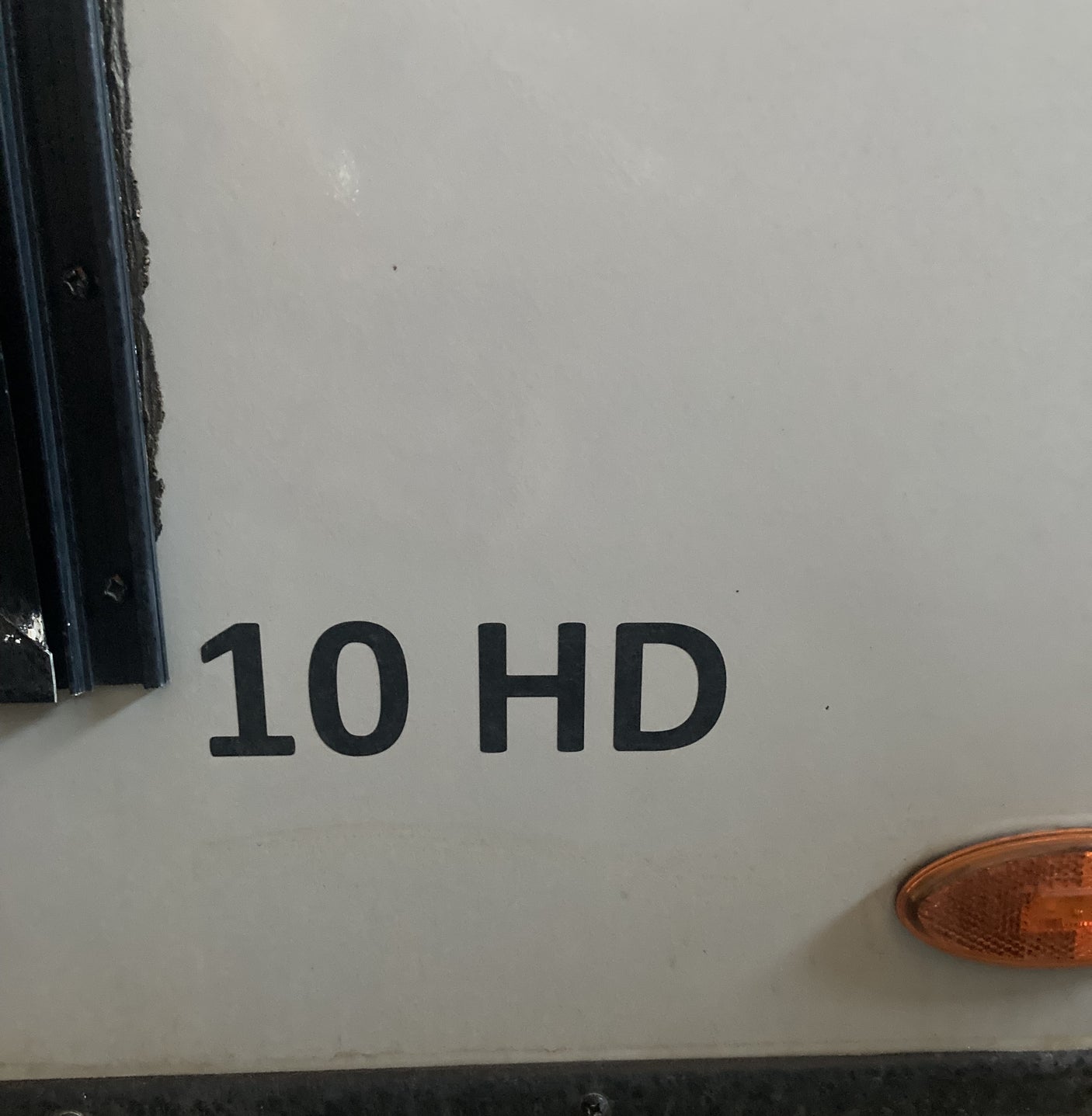 Close-up of an RV rental exterior. Black '10 HD' lettering on light gray, with black trim on the left and an amber light on the right.
