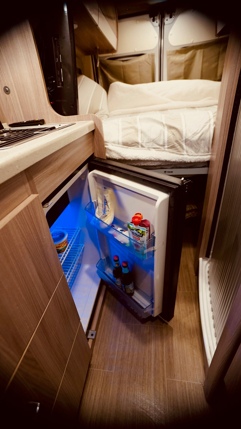 Inside your RV rental: a compact kitchen with an open, blue-lit fridge, and a cozy bed beyond. Perfect for travel. — photo 7