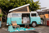 Stylish blue & white VW camper van, an ideal RV rental, open to show its cozy interior, with a dog, chair, and rug outside.
