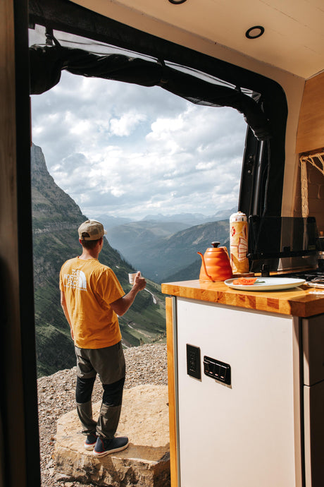 RV rental: Man sips coffee from his camper van, overlooking a stunning mountain panorama.