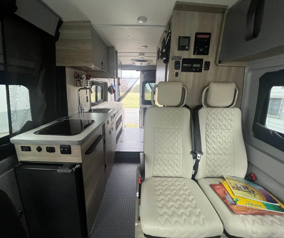 Inside a modern RV rental camper van. Features a kitchen with stove & sink, light seats, and control panels. Open rear exit.