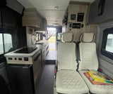 Inside a modern RV rental camper van. Features a kitchen with stove & sink, light seats, and control panels. Open rear exit.