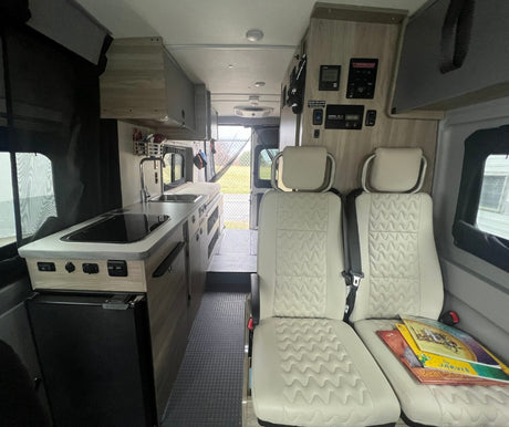 Inside a modern RV rental camper van. Features a kitchen with stove & sink, light seats, and control panels. Open rear exit.