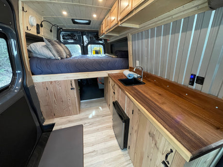 Cozy **rv rental** camper van interior featuring an elevated bed, compact kitchen with sink, fridge, and warm wooden accents.