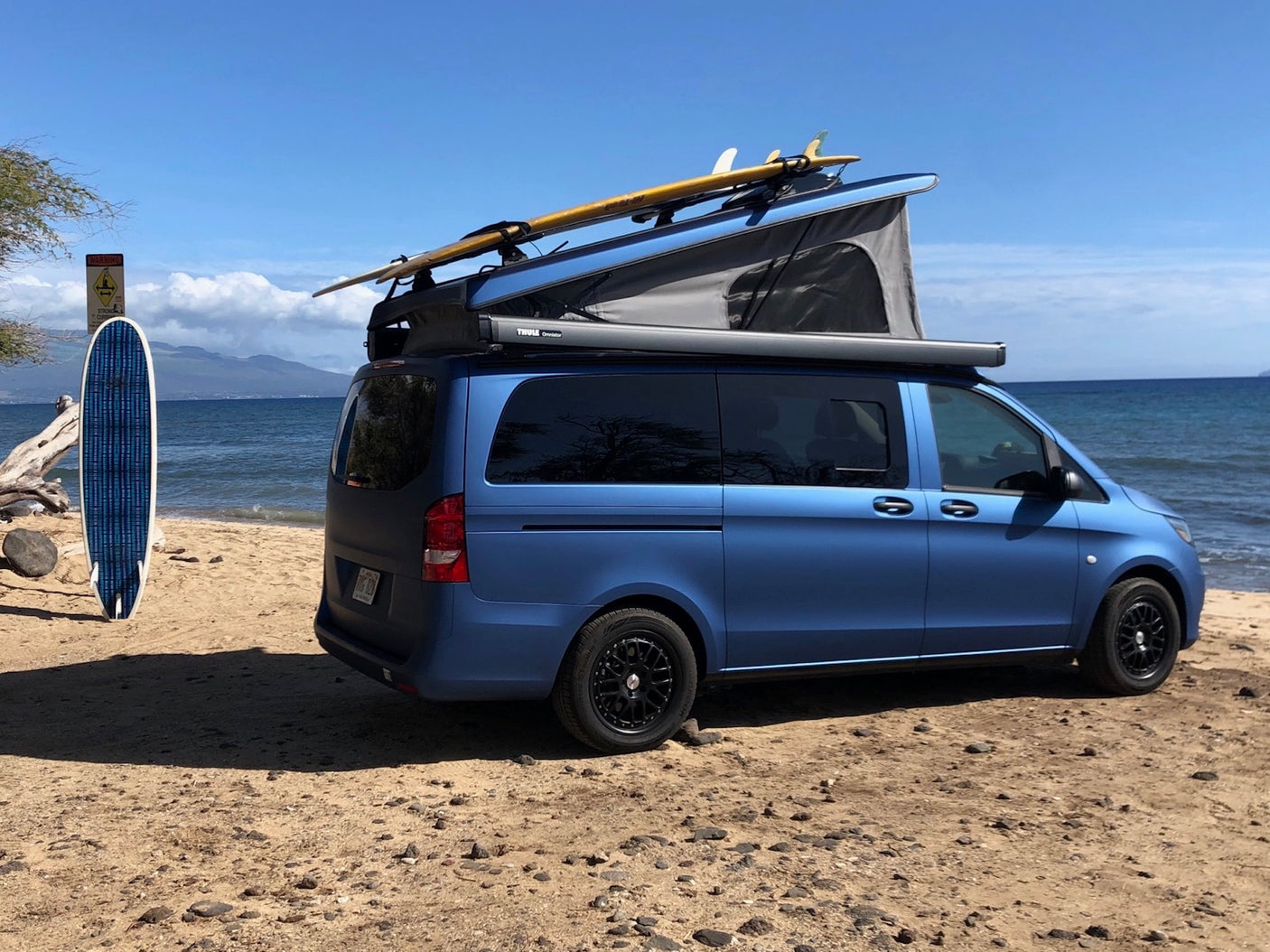 A blue pop-top camper van with surfboards on a sandy beach. Ideal RV rental for your next beach and surf adventure!