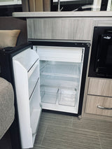 Open mini-fridge with empty shelves & drawers, next to a microwave in a clean RV rental kitchenette.