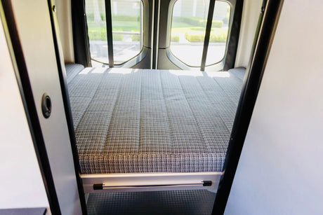Inside this rv rental, a comfy campervan bed with a patterned mattress and rear windows, ready for your next adventure.