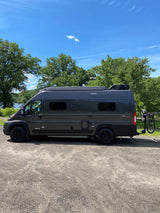 This gray Winnebago Solis RV rental van, with a bike rack, is parked on gravel near a lake and trees.