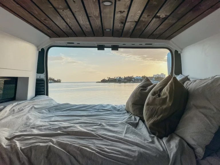 Cozy RV rental bed inside a van, overlooking a serene ocean sunset. Enjoy scenic travel and van life adventures!
