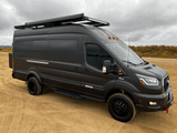 Black custom Ford Transit camper van, ideal for rv rental. Features off-road tires, roof rack on sandy terrain.