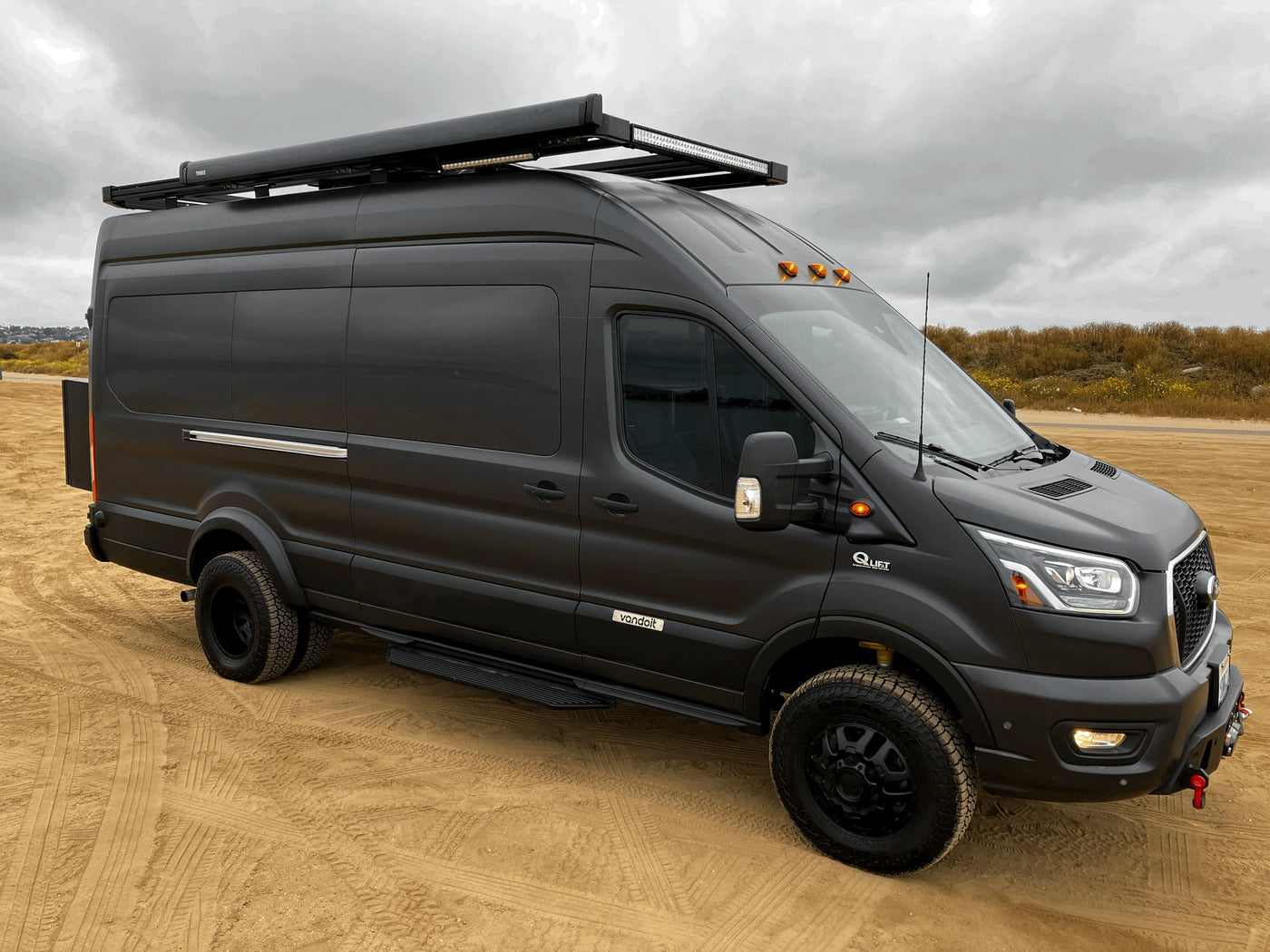 Black custom Ford Transit camper van, ideal for rv rental. Features off-road tires, roof rack on sandy terrain. — photo 4