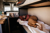 Comfortable RV rental interior: A person sleeps soundly on a lower bunk, with another bed and kitchen area visible, offering cozy van life travel.
