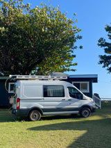 White Ford Transit rv rental camper van parked on green grass. A house and large tree are behind it under a blue sky.