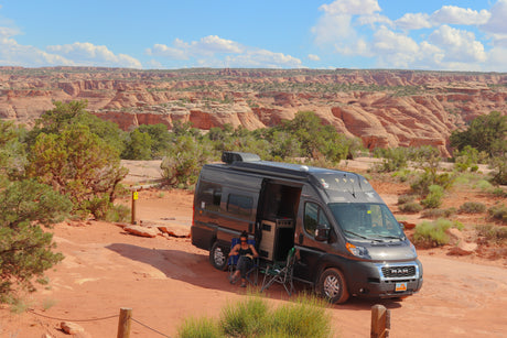 A person reads in an RV rental camper van parked at a scenic red rock canyon campsite, offering stunning desert views.