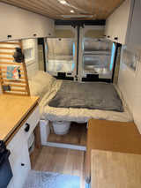 Inside a cozy rv rental camper van: bed at rear, compact kitchen on left, overhead cabinets, and wood ceiling.