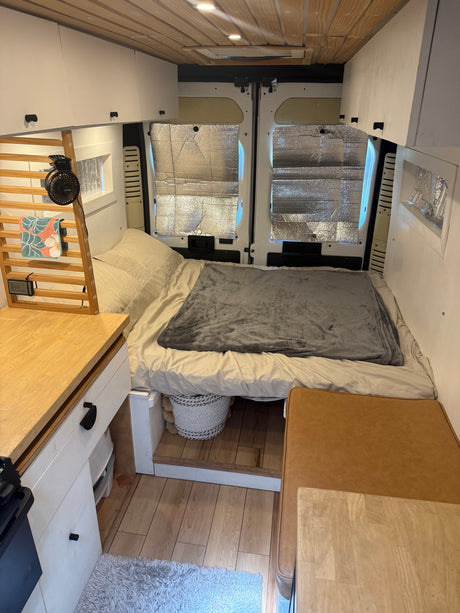 Inside a cozy rv rental camper van: bed at rear, compact kitchen on left, overhead cabinets, and wood ceiling.