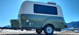 This compact, retro-style RV rental trailer, with utility hookups and rooftop AC, is parked against a backdrop of snowy mountains.