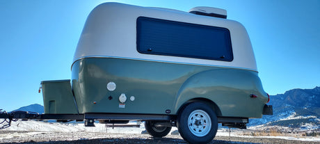 This compact, retro-style RV rental trailer, with utility hookups and rooftop AC, is parked against a backdrop of snowy mountains.