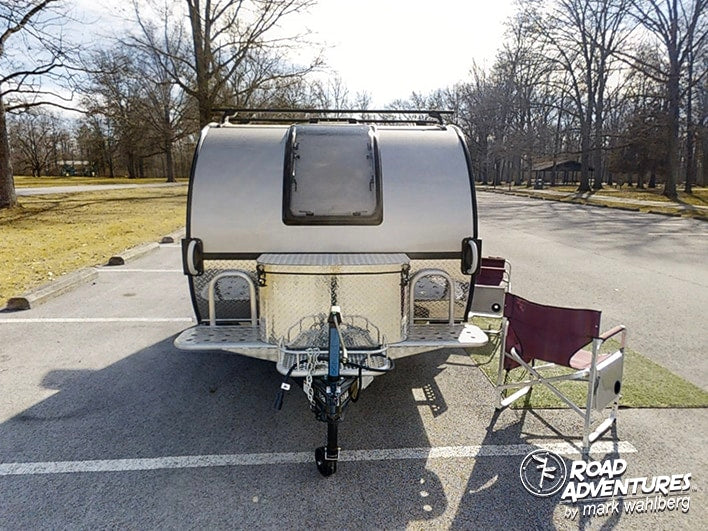 This silver teardrop RV rental is parked, ready for camping. A chair sits nearby in a scenic park setting.